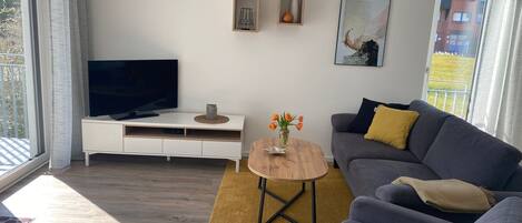 Una sala de estar moderna con un televisor de pantalla plana sobre una consola blanca, un sofá gris con un cojín amarillo, una mesa de centro de madera con un jarrón de flores, y un cuadro abstracto enmarcado en la pared.