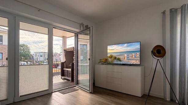 Una sala de estar moderna con un televisor grande, una lámpara de pie y un amplio ventanal corredera que da a un balcón.