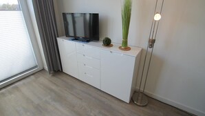 Un salon moderne avec un téléviseur à écran plat posé sur un meuble blanc, une petite plante et une lampe sur pied.