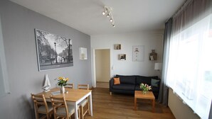 Ein Wohnzimmer mit einem Holzesstisch, Stühlen, einem schwarzen Sofa und einem Couchtisch. An der Wand hängt ein gerahmtes Bild und es gibt ein Fenster mit Vorhängen.