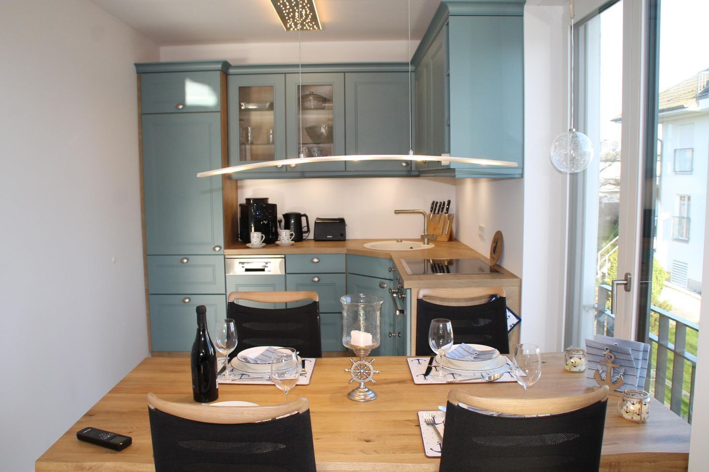 A modern kitchen with light blue cabinets, a wooden dining table set for four, and a large window providing a view of the outside.