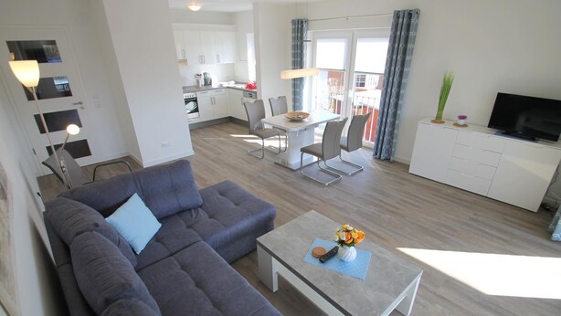 A modern living room with a gray sectional sofa, a coffee table with a vase of flowers, and a dining area with a white table and chairs. - 2104 - Strandwiese - 2104 - Strandwiese 9 - Apartment 4 (Dahme)