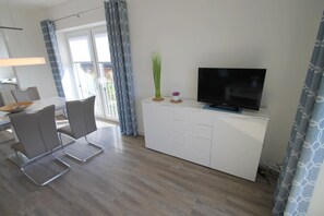 Un salon moderne avec un téléviseur à écran plat posé sur un meuble blanc, des fauteuils gris et une table à manger agrémentée d’un panier.