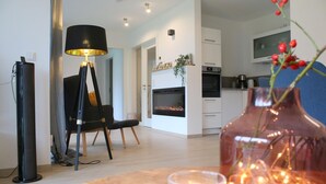 A modern living room with a fireplace, a dining area with a table and chairs, and a kitchen with white cabinets and a built-in oven. - 2602 - Strandwiese 18 - Apartment 2602 in the Strandwiese (Dahme)