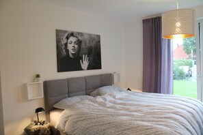 Et soveværelse med en stor seng, en grå sengegavl, et billed ophængt på væggen, en senelampe og et vindue med gardiner.