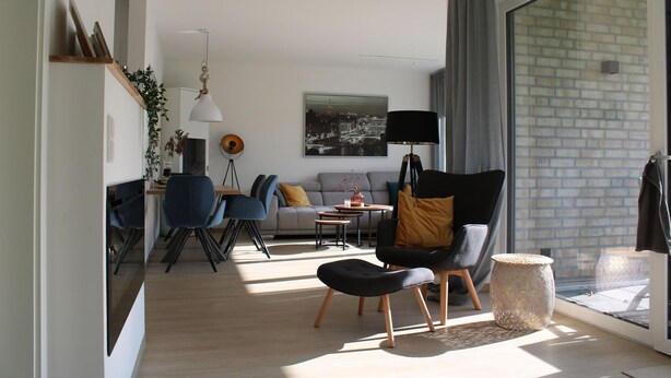 A modern living room with a sofa, chairs, a dining area, and a large window with a view of a brick wall. - 2602 - Strandwiese 18 - Apartment 2602 in the Strandwiese (Dahme)