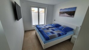 Ein Schlafzimmer mit einem Bett, einem Fenster, einem an der Wand montierten Fernseher und einem Bild.
