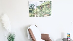 A room with a brown chair, a white fluffy cushion, a potted plant, and a wall-mounted picture of a Hobbiton-like house.