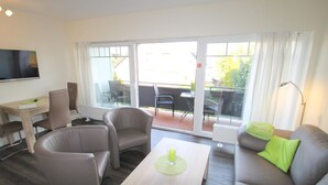 Una sala de estar moderna con un sofá, dos sillones, una mesa de centro y un televisor. Hay una puerta corrediza de vidrio que da a un patio exterior con sillas y una mesa.
