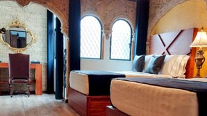 Un dormitorio con cama de madera, un escritorio y una silla. Hay dos ventanas de arco y una araña.