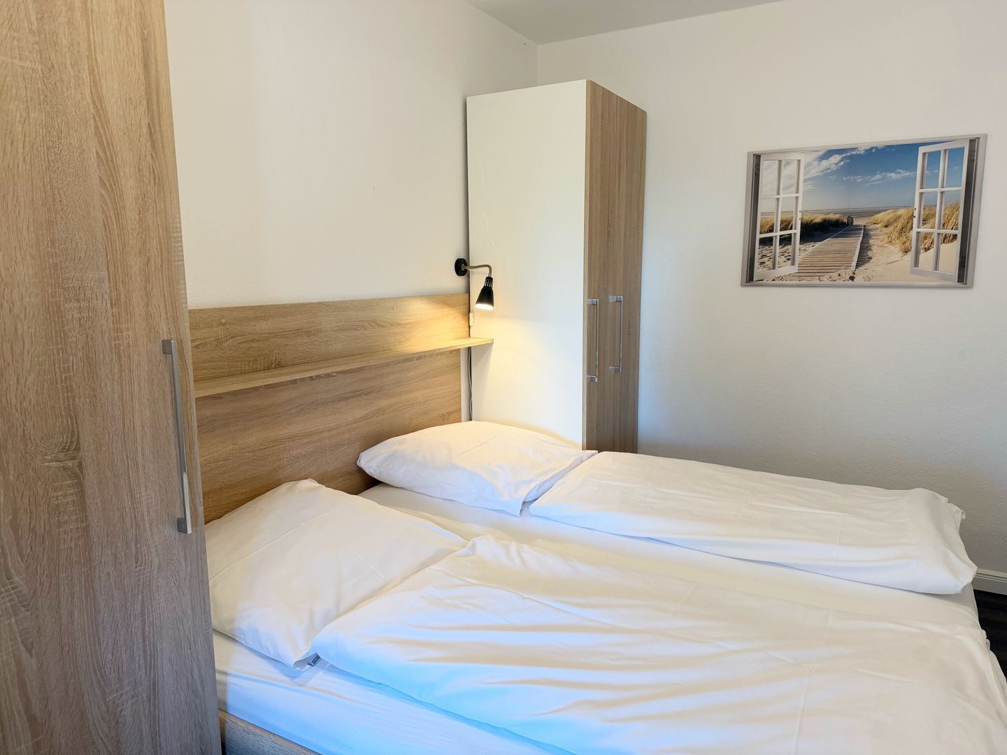 Een slaapkamer met een houten hoofdbord, een bedlampje, een bed met wit beddengoed, een kledingkast en een wanddecoratie met een strandthema.