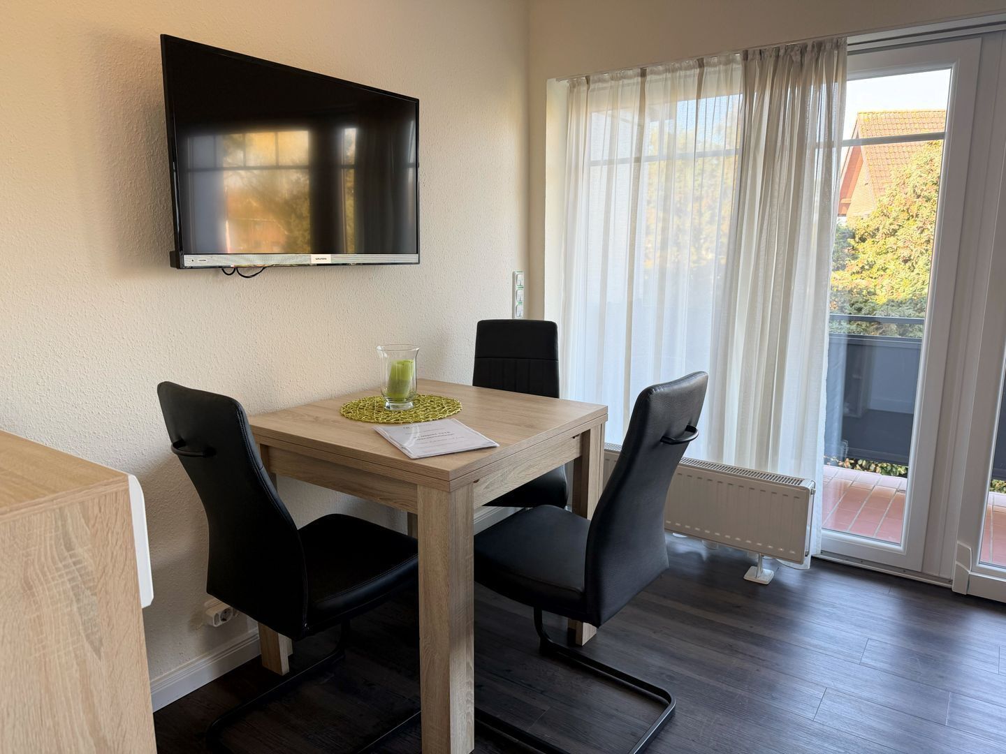 Ein Zimmer mit Holztisch, schwarzen Stühlen, einem an der Wand befestigten Flachbildfernseher und einem Fenster mit durchsichtigen Vorhängen.
