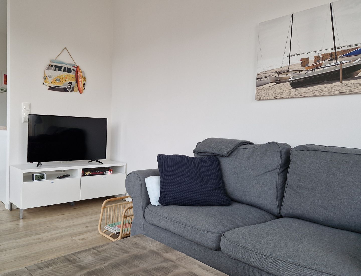 Un salon avec un canapé gris, un téléviseur à écran plat posé sur un meuble blanc, un panier en osier et un tableau représentant des bateaux accroché au mur.