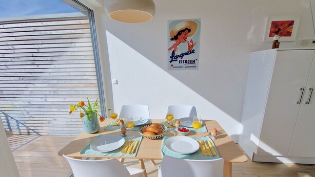 Un coin repas avec une table dressée pour deux personnes, comprenant des assiettes, des couverts et un panier de pain. Une affiche est accrochée au mur, et on aperçoit un meuble en arrière-plan.