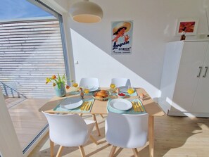 Zona pranzo con tavolo apparecchiato per due, piatti, posate e una ciotola di pane. Una locandina è appesa al muro, e si intravede un mobile sullo sfondo.