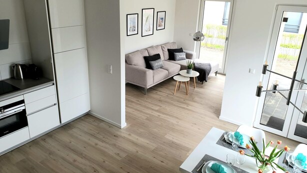 Une cuisine moderne avec des meubles blancs, un coin repas avec une table dressée pour deux personnes, et un salon avec un canapé et une table basse.