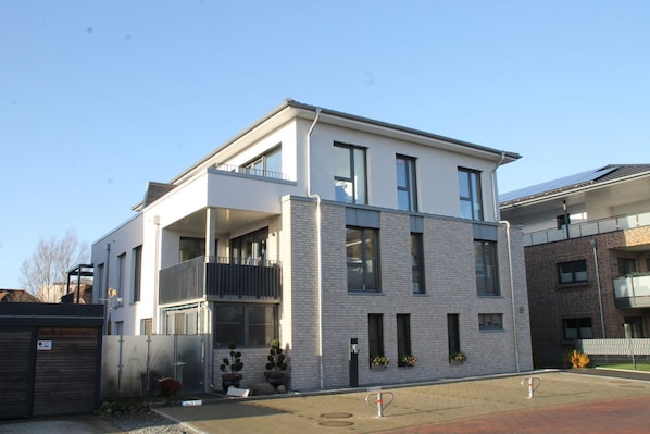 A modern two-story residential building with a balcony on the upper floor, a garage, and a small garden area with potted plants.