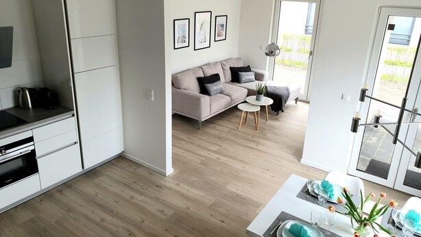A modern kitchen with white cabinets, a dining area with a table set for two, and a living room with a sofa and a coffee table. - 2401 - Strandwiese - Apartment 2401 in the Strandwiese (Dahme)