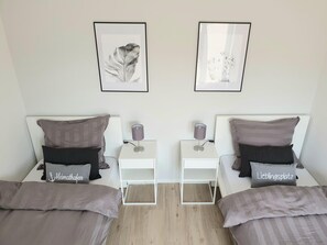 A twin room with two beds, each with a pillow labeled 'Heimathafen' and 'Lieblingsplatz,' a white nightstand with a lamp, and two framed pictures on the wall.