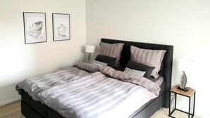 Un lit avec une tête de lit sombre, une table de chevet avec une lampe, et deux tableaux encadrés accrochés au mur.