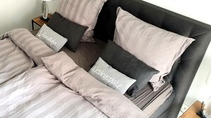Un letto rifatto con cura, con lenzuola a righe e cuscini con scritto 'Heinathof' e 'Lieblingplatz'.