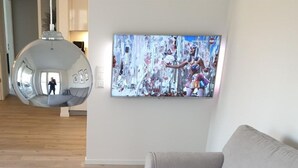 Un salon avec un canapé gris, une table basse blanche ornée d’un vase de fleurs, un grand téléviseur à écran plat fixé au mur et un luminaire suspendu.