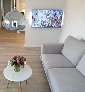 Een woonkamer met een grijze bank, een witte salontafel met een bloevas, een grote flatscreen televisie aan de muur en een hanglamp.