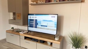 Una sala de estar moderna con un televisor mostrando un sitio web, un mueble de madera con espacio de almacenamiento, una estantería con objetos decorativos y una planta en maceta.
