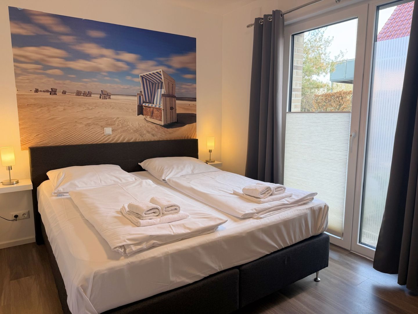 A hotel room with a bed, two bedside tables, a window with curtains, and a beach-themed wall mural.