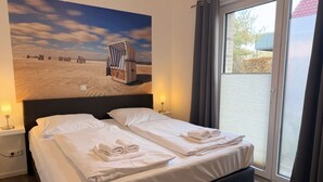 Una habitación de hotel con cama, dos mesitas de noche, una ventana con cortinas y un mural en la pared con temática playera.
