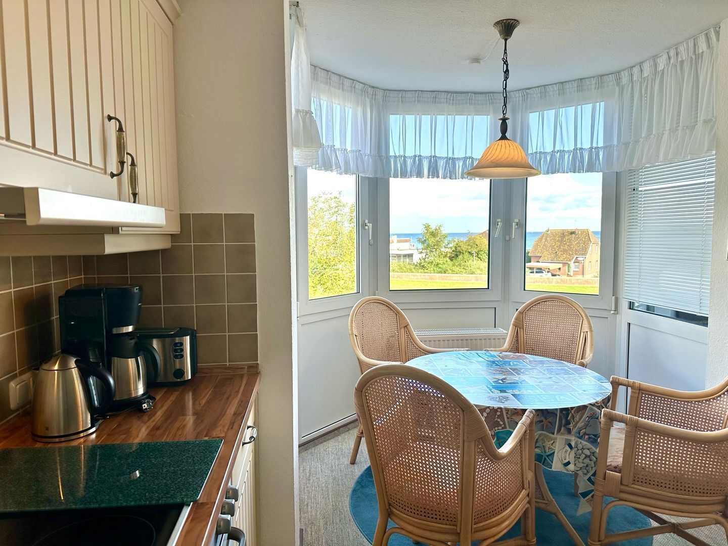 A kitchen with a coffee maker, a toaster, and a kettle. There is a dining table with four wicker chairs and a blue tablecloth. A window with a view of the sea and a building is visible.