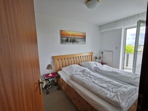 Et soveværelse med en træseng, et natbord med lampe, en radiator og et vindue med udsigt over grønne omgivelser.