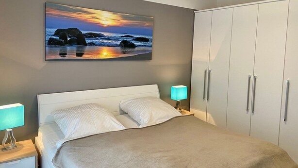 A bedroom with a bed, a nightstand, a wardrobe, and a sunset painting on the wall.