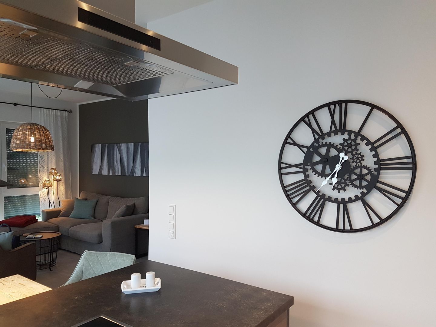 A modern living room with a large wall clock, a dining area with a dark table, and a kitchen with a range hood.