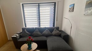 Una sala de estar moderna con un sofá seccional gris, una pequeña mesa redonda de madera con un florero de flores rojas y un amplio ventanal con persianas.