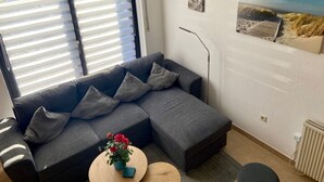 Una sala de estar moderna con un sofá seccional gris, una pequeña mesa redonda de centro con un jarrón de flores rojas y un ventanal con persianas.