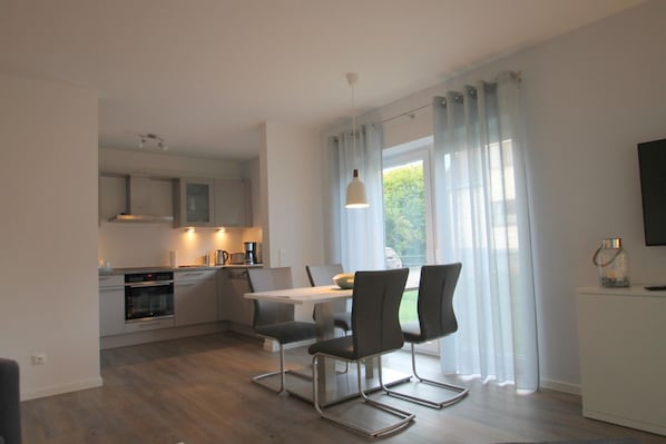 A modern kitchen with a dining area, featuring a table with chairs, a TV on a stand, and a view through large windows.