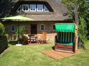 A house with a thatched roof, a wooden deck, a green umbrella, and a wicker chair.