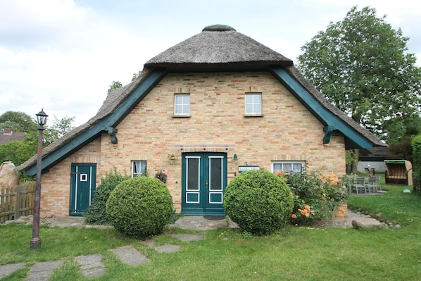 A stone cottage with a thatched roof, two green doors, and a small garden with a bench and flowers.