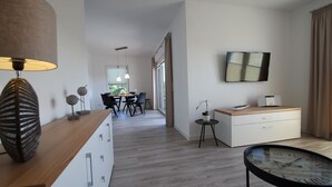 A modern hotel room with a wooden desk, a dining area with a table and chairs, a white dresser, a clock, and a TV mounted on the wall.