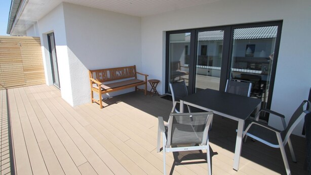 A covered outdoor area with a wooden bench, a table with chairs, and a glass door leading to an interior space.