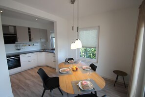 Une cuisine moderne avec des meubles blancs, un four encastré et un coin repas comprenant une table en bois et des chaises noires.