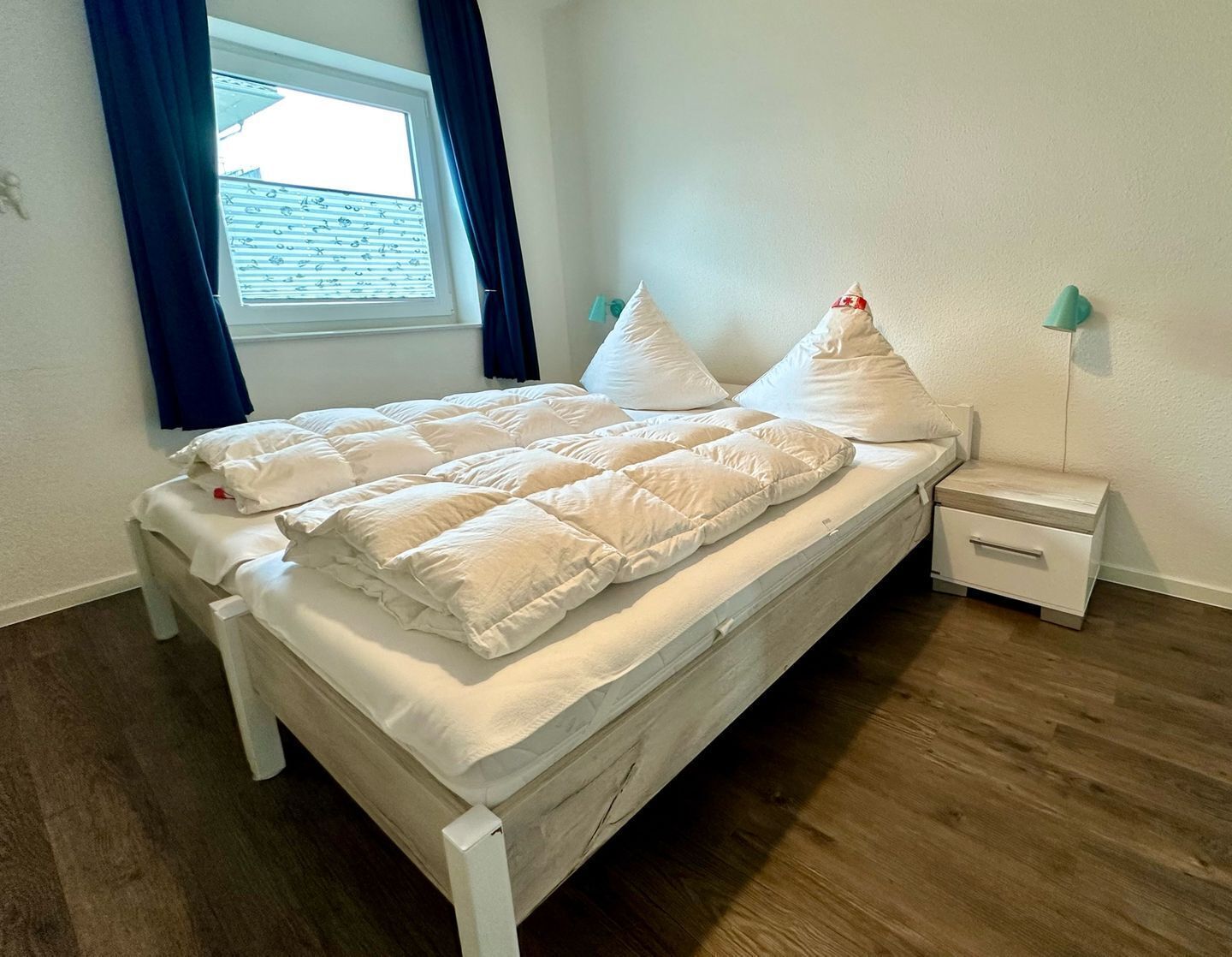 Uma cama bem arrumada com roupa de cama e almofadas brancas, piso de madeira, janela com cortinas azuis e mesa de cabeceira com abajur.