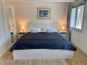 A neatly made bed with a dark quilt, two white pillows, and a framed picture above it. There are two bedside tables with lamps, and a window with curtains on the right.