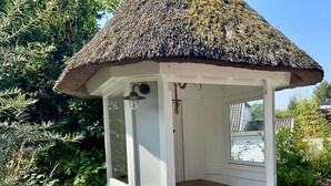 A wooden gazebo with a thatched roof, a bench inside, and a view of the surrounding greenery.