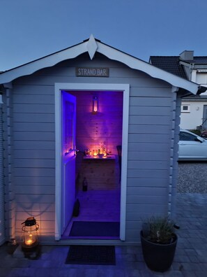 A small wooden structure labeled "STRAND BAR" with a welcoming entrance, a small table with bottles, and a lantern outside.