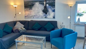 A living room with a grey sofa, a blue armchair, a glass coffee table, and a painting of a lighthouse on the wall. - 1805 - Ostseestern - 1805 - Ostseestern in Dahme (Dahme)