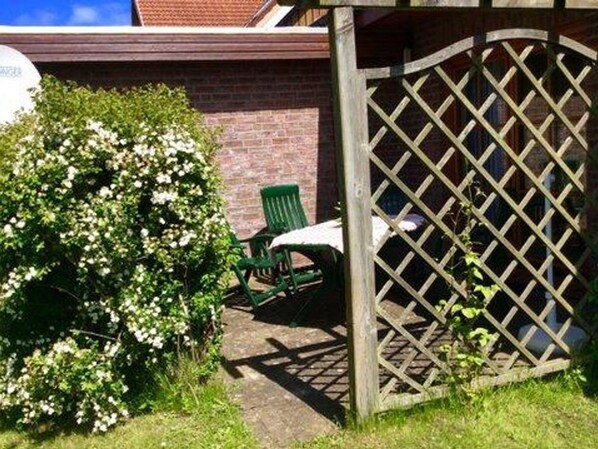 Un jardín con un portón de madera con celosía, una silla verde y un arbusto de flores blancas.