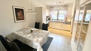 A compact kitchen with white cabinets, a microwave, and a sink. There's a dining area with a table and chairs, and a window with curtains. - 2050 - House Heisterbusch (Kellenhusen (Ostsee))