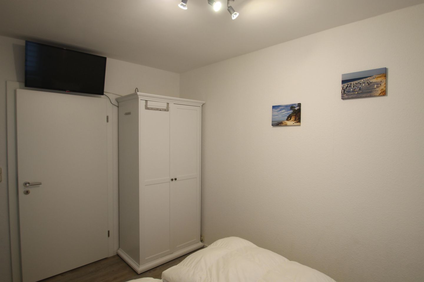 A bedroom with a bed, a wardrobe, a television on the wall, and two framed pictures.